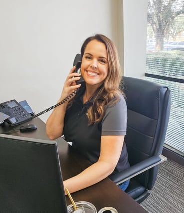 Smiling woman in black polo shirt on office phone, seated at desk with computer.