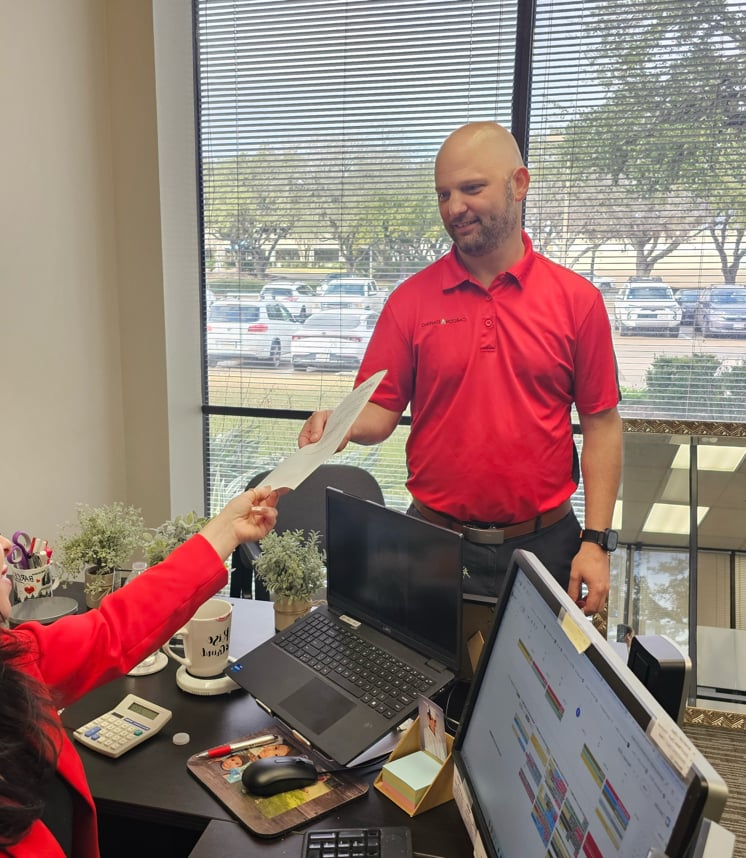 Man in red shirt hands paper to woman in red jacket at office desk with laptops.