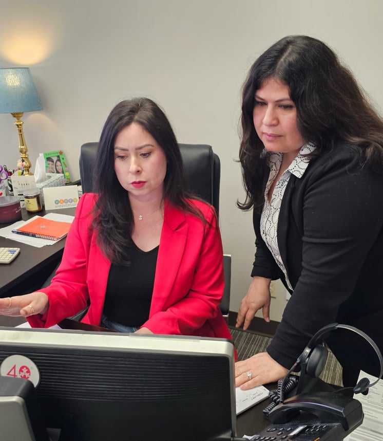 Two women in an office discussing work at a computer, one in a red blazer.